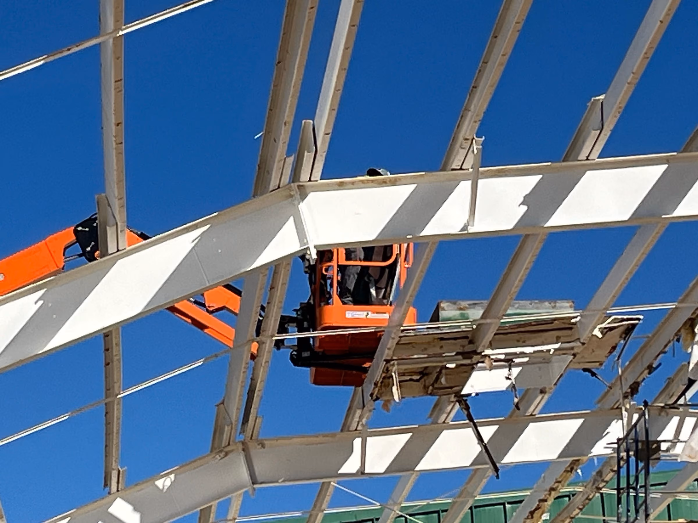 A worker operating a boom lift from the basket among structural steel roof framing